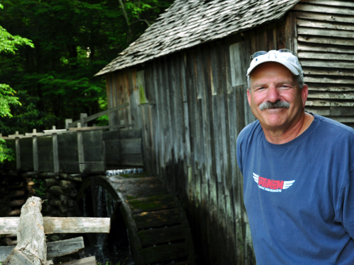 Man wearing a white baseball cap standing next to a wood shed